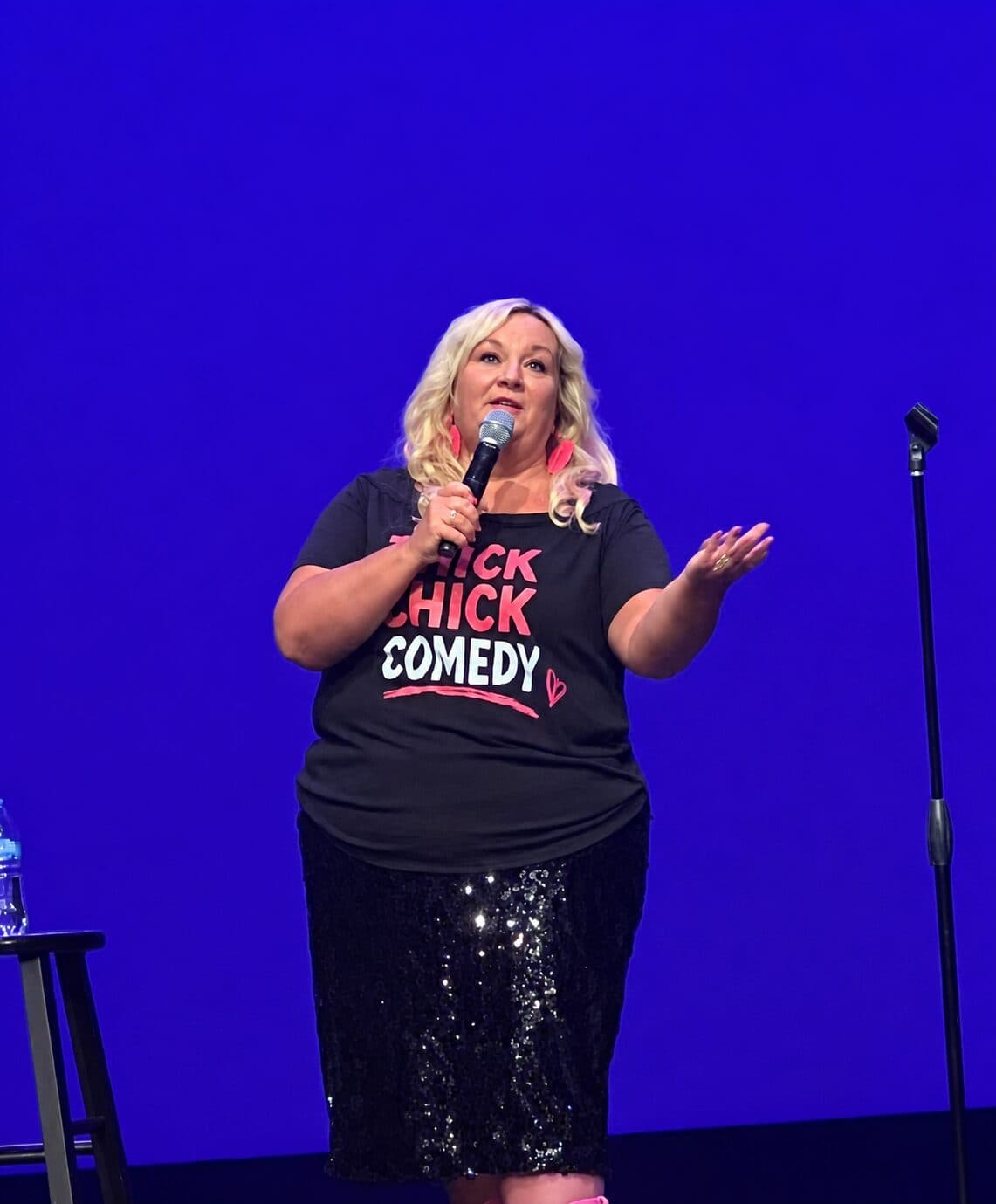 Susan Horne on stage mid-punchline in her Thick Chick Comedy T-shirt, microphone in hand and pink boots peeking out below a sequined skirt